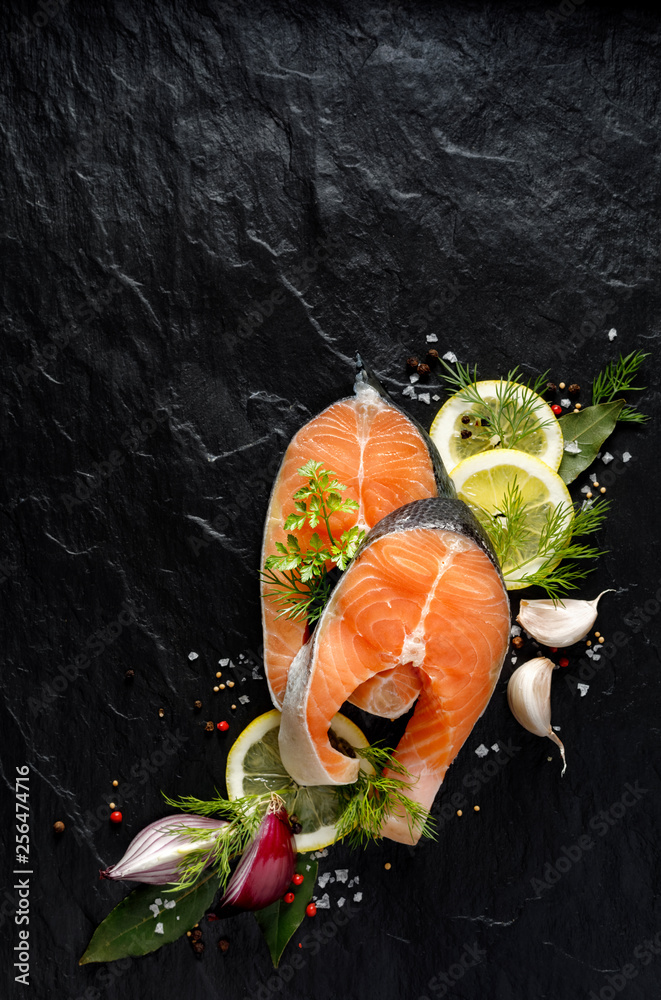 Φωτογραφία Fresh, raw salmon steaks with the addition of slices of lemon, garlic, onion, herbs and spices on a black stone background, top view