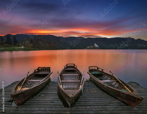 Fototapeta Naklejka Na Ścianę i Meble -  Bled, Slovenia - Traditional boats at Lake Bled with beautiful dramatic sunset and the Assumption of Mary Pilgrimage Church and mountains at background at sunset