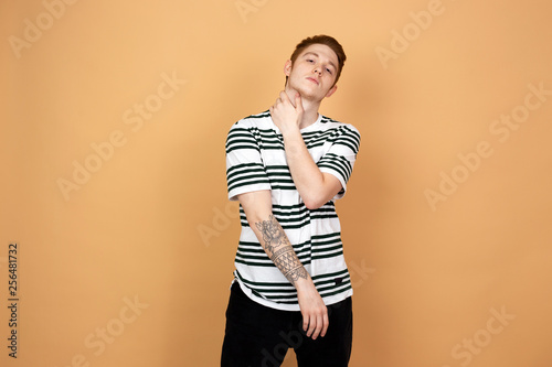 Red-haired stylish guy in a striped shirt and black jeans with tattoo on his hand stands on the beige background in the studio