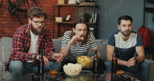 Young Caucasian guys watching sport game attentively on TV while sitting in the cozy living room and eating chips and popcorns with beer. photo