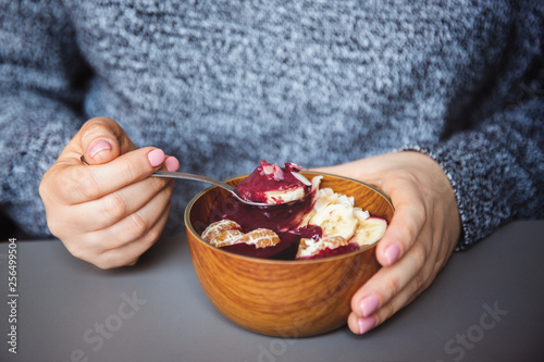 Acai smoothie, granola, seeds, fresh fruits in a wooden bowl in female hands on grey table. Eating healthy breakfast bowl photo