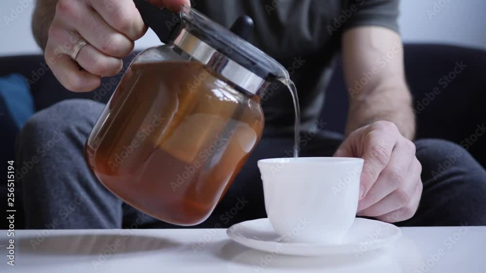 A man at home pours tea from a glass teapot into a porcelain white cup