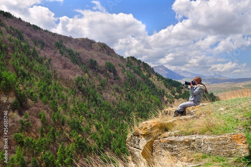 Girl sitting on the edge of a cliff and taking pictures of the mountain landscape