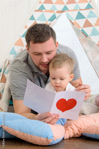 Happy father with his one-year-old son, reading a gift card with a red heart. Father day, valentines day.