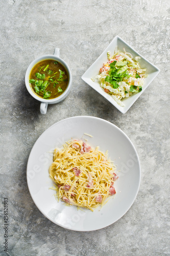 Business lunch menu, pasta Carbonara, green salad and chicken soup. Gray background, top view, space for text