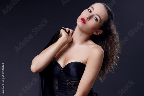 Portrait of beautiful young girl in a silk black dress with lace corsage on dark background. Elegant woman in a black dress on a dark background