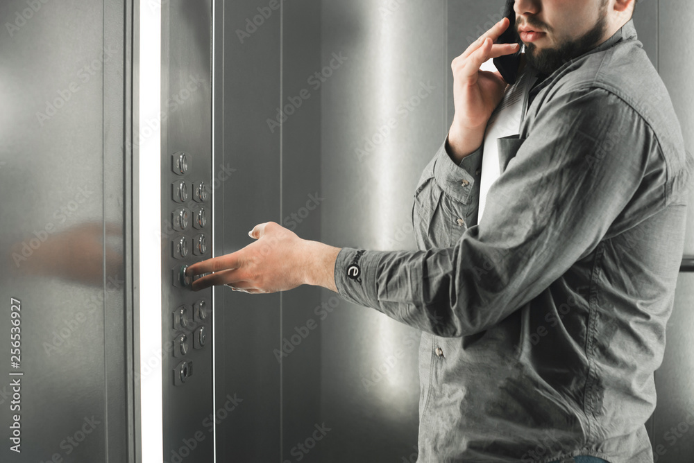 Diligent businessman pressing button inside elevator and talking on the phone. Man office worker in a grey shirt is pushing the button with her forefinger. Diligent businessman pressing button inside elevator and talking on the phone. Man office worker in a grey shirt is pushing the button with her forefinger.