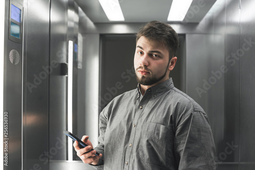 Serious bearded man in a shirt stands in the elevator with a smartphone in his hand and is concentrated in the camera. Portrait of a serious business man in the elevator.
