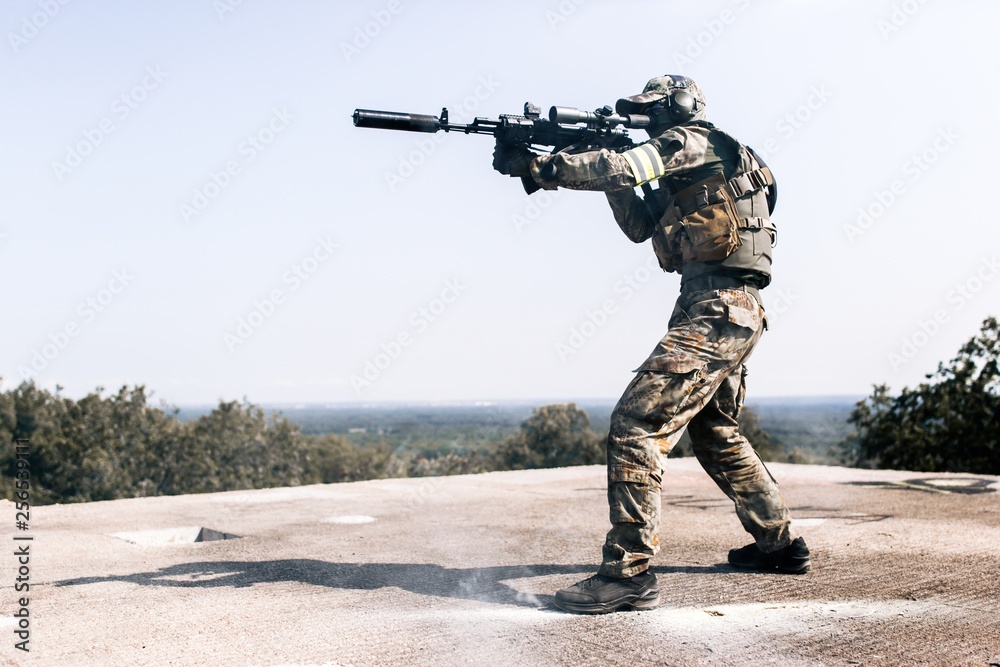 Fototapeta premium Armed man in camouflage with sniper gun in hand standing on the roof