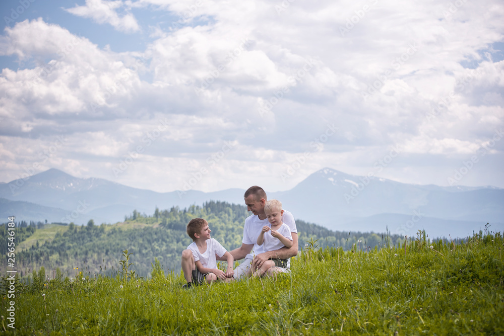 Happy father with his two young sons sitting on the grass on a background of green forest, mountains and sky with clouds. Friendship concept.