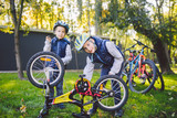 Two children, older boys and younger brother learning repair bike. Two guys siblings in helmets and single clothes use pump tool and pump air into bicycle wheel in park background of the house
