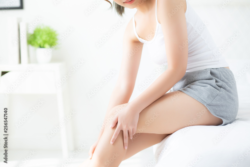 Obraz premium Beautiful young asian woman sitting on a bed stroking legs with soft smooth skin in the bedroom, girl applying body cream and lotion with treatment care, healthy and wellness concept.