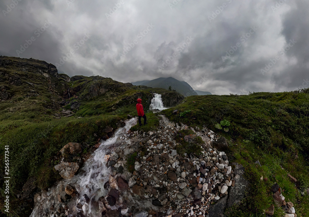 man in red stands under waterfall on black rock in mountains on cloudy rainy day.. Wide panorama