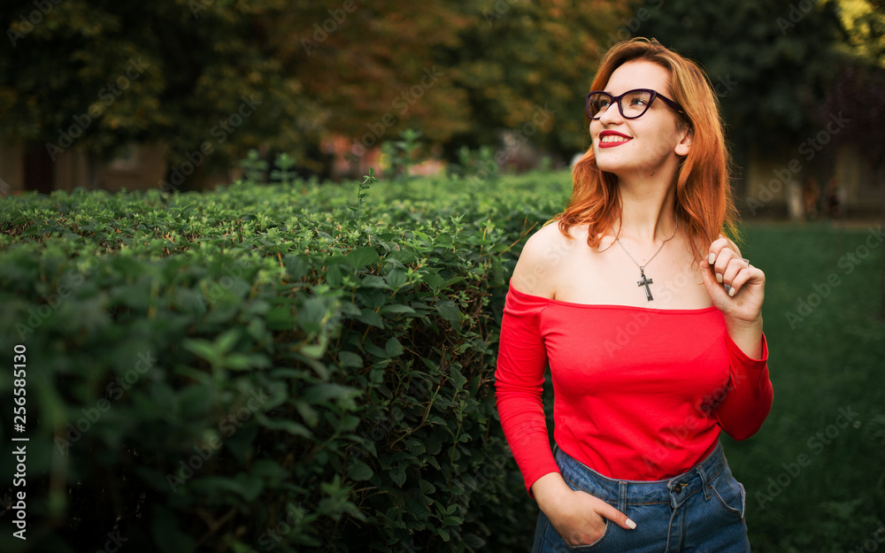 Attractive redhaired woman in eyeglasses, wear on red blouse and jeans skirt posing at green park. Attractive redhaired woman in eyeglasses, wear on red blouse and jeans skirt posing at green park.