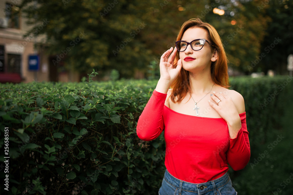 Attractive redhaired woman in eyeglasses, wear on red blouse and jeans skirt posing at green park. Attractive redhaired woman in eyeglasses, wear on red blouse and jeans skirt posing at green park.