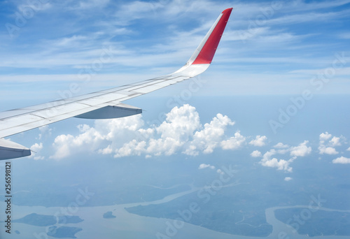 Wing of an airplane flying above clouds and river mouth scenery below as background.