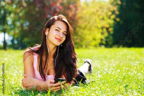 Young woman in dress lies on green grass and listens to music in headphones through smartphone.