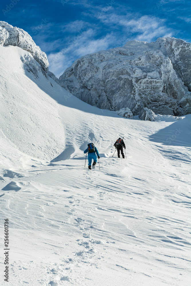 A trip to the mountains. Two tourists walk up a snowy slope under icy rocks on a sunny day.
