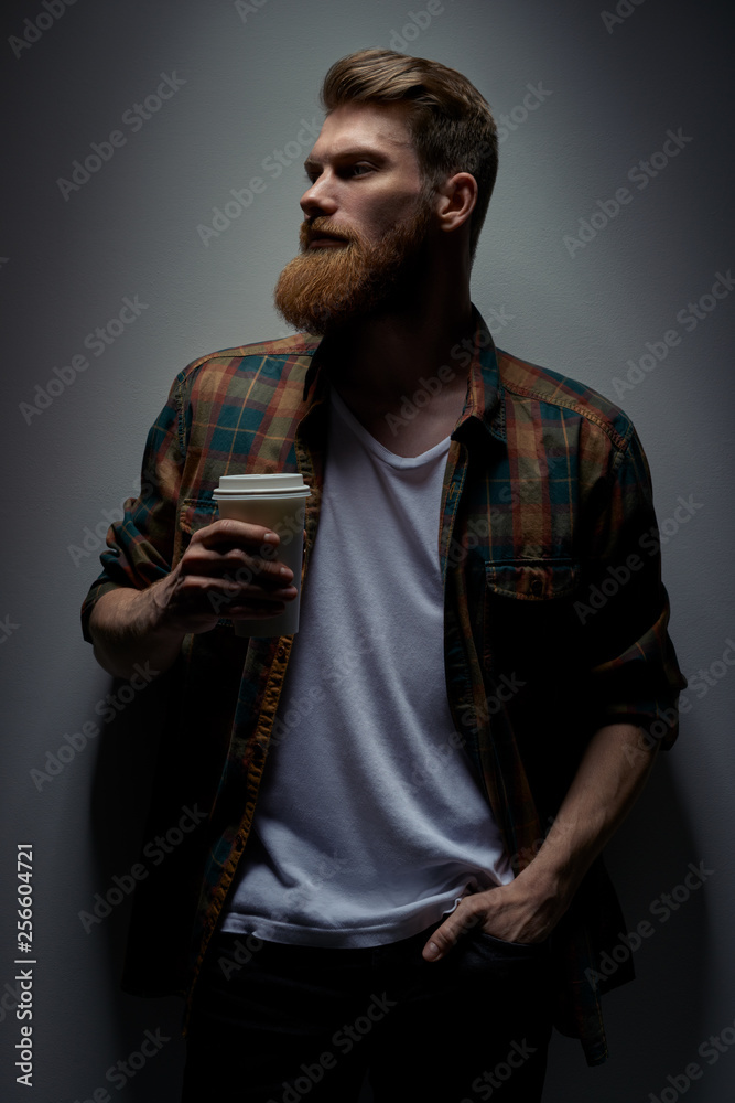 Naklejka premium Side view shot of young bearded stylish hipster with coffee in hand Handsome confident perfect hairstyle man having coffee indoor Male taking rest drinking fresh morning coffee to go.