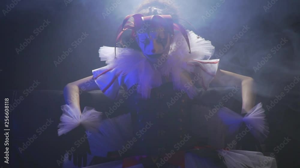 Creepy clown with white and black makeup is wearing a dress and sitting in the smoke under the spotlight Creepy clown with white and black makeup is wearing a dress and sitting in the smoke under the spotlight