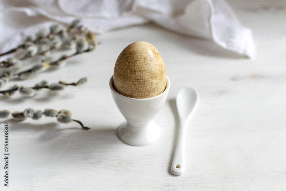 Fototapeta premium Naturally dyed easter egg for breakfast, white egg cup, draped napkin, white wooden table, willow banch - minimal easter concept
