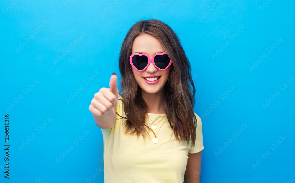 summer, valentine's day and people concept - smiling young woman or teenage girl in yellow t-shirt and heart-shaped sunglasses showing thumbs up over bright blue background
