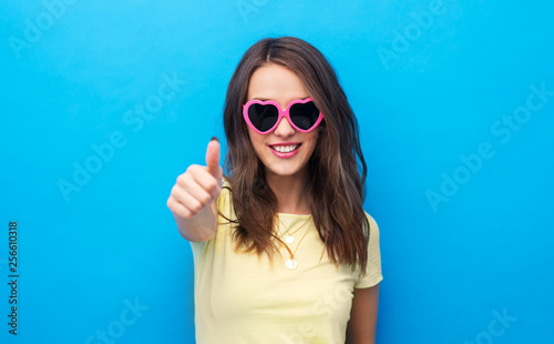 summer, valentine's day and people concept - smiling young woman or teenage girl in yellow t-shirt and heart-shaped sunglasses showing thumbs up over bright blue background