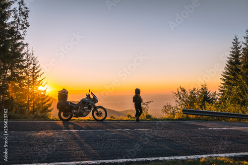 girl in full motorcycle equipment, stands on the side of the road over a cliff and looks into the distance at beautiful sunset in the mountains. Adventure motorcycle, Transfagarasan, Romania © Sergey