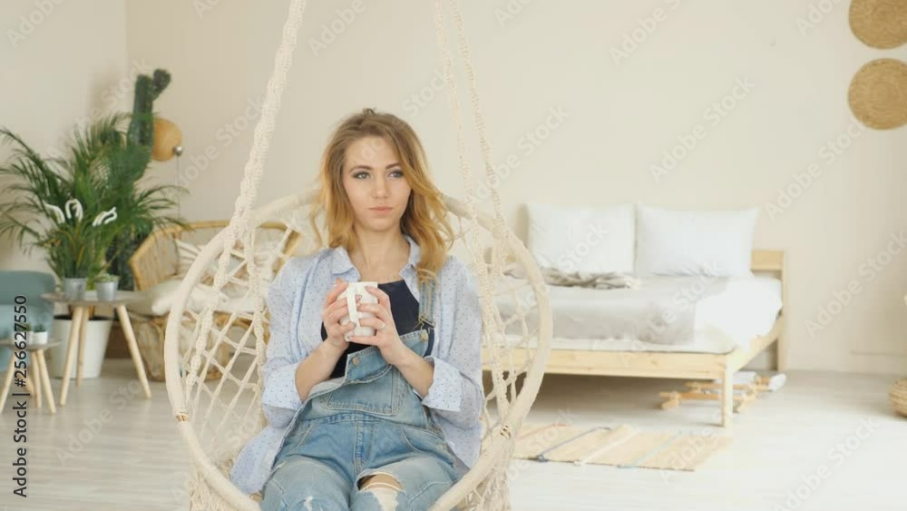 pretty blond girl with long hair wearing jeans sits in hanging wicker chair drinking coffee in modern light bedroom