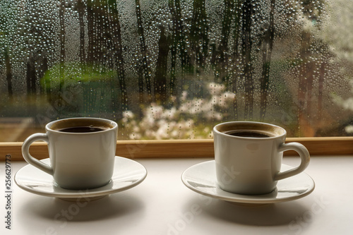 Two hot coffee cups on a rainy spring morning against a window background covered in raindrops with blurred cherry blossom trees.