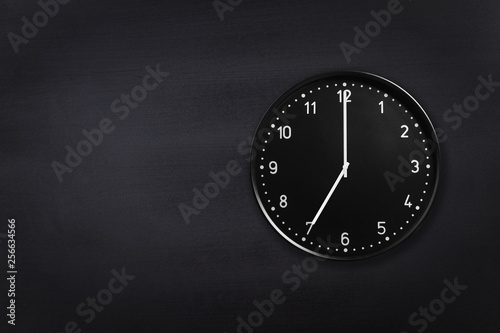 Black wall clock showing seven o'clock on black chalkboard background. Office clock showing 7am or 7pm on black texture photo