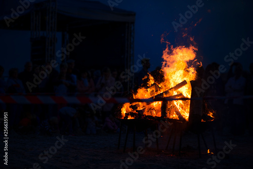 Large burning bonfire with soft glowing flame and sparkles flying all around. Romantic summer evening, people relaxing and enjoying calmness at the festival on seaside. Stage, singers in background © lainen