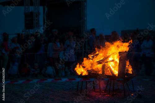 Large burning bonfire with soft glowing flame and sparkles flying all around. Romantic summer evening, people relaxing and enjoying calmness at the festival on seaside. Stage, singers in background © lainen