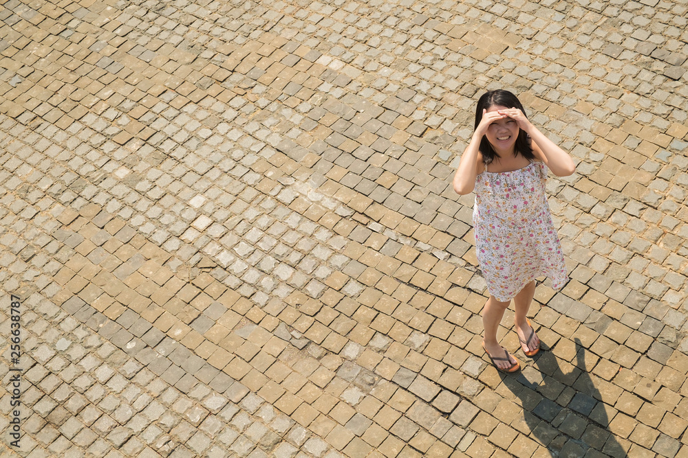 Asian standing on brick ground in harsh sun light with her hands try to block the sun
