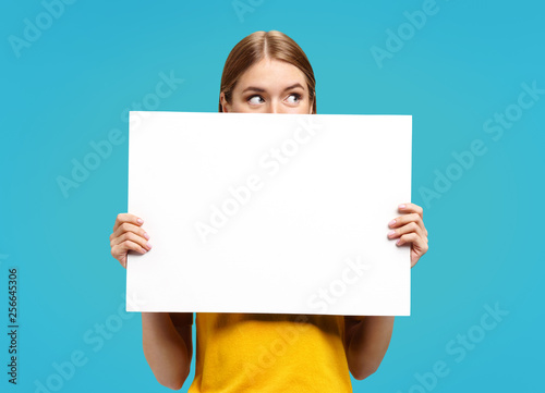 Funny girl with white empty poster, looking away, on blue background. Copy space for your text. photo