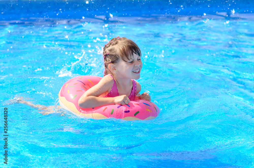 Cute smiling little girl with inflatable donut circle learns to swim in swimming pool on hot sunny day. Healthy and happy childhood concept.