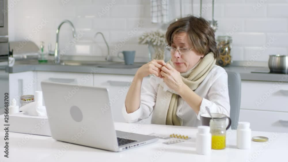 Medium shot of middle-aged Caucasian woman sitting at laptop, coughing and sneezing and talking with friend by internet Medium shot of middle-aged Caucasian woman sitting at laptop, coughing and sneezing and talking with friend by internet