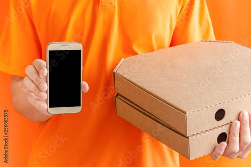 Delivery man in orange shirt holding stack of pizza boxes and showing copyspace screen of cell phone. concept call, text, applications isolated over black background