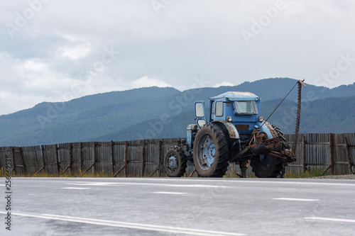 Blue old tractor with big wheels on the side of the road next to a wooden fence.  Mower device. the old horse mower. Mountain landscape on a cloudy day