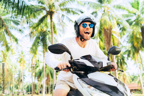 Happy smiling and screaming male tourist in helmet and sunglasses riding motorbike scooter during his tropical vacation under palm trees.