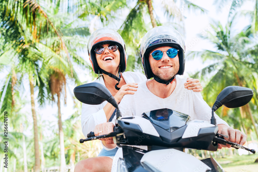 Happy smiling couple travelers riding motorbike scooter in safety helmets during tropical vacation under palm trees - obrazy, fototapety, plakaty 