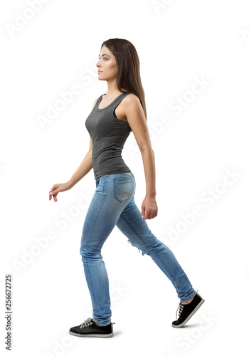 Side view of young woman in gray sleeveless top and blue jeans, with long dark hazelnut hair, walking forward on white background.