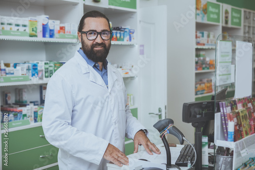 Wallpaper Mural Cheerful mature male druggist smiling joyfully to the camera, standing behind checkout counter at his drugstore. Happy bearded chemist enjoying working at pharmacy. Medicine, sales concept Torontodigital.ca