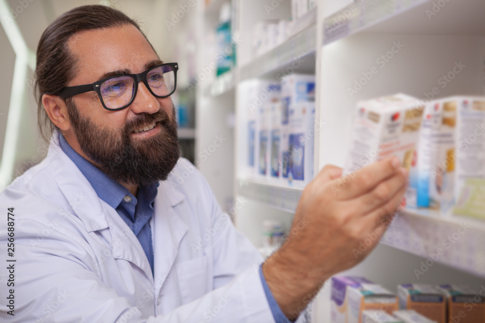 Close up of a cheerful handsome bearded pharmacist smiling, working at the drugstore, copy space. Happy male chemist organizing medical goods for sale. Retail, consumption concept Close up of a cheerful handsome bearded pharmacist smiling, working at the drugstore, copy space. Happy male chemist organizing medical goods for sale. Retail, consumption concept