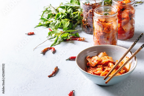 Korean traditional fermented appetizer kimchi cabbage and radish salad, hot spicy anchovies fish snack served in glass jars and bowl with Vietnamese oregano and chili peppers over grey blue table. photo