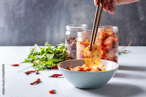Korean traditional fermented appetizer kimchi cabbage and radish salad, fish snack served in glass jars with Vietnamese oregano and chili peppers over grey blue table. Chopsticks in mans hands.