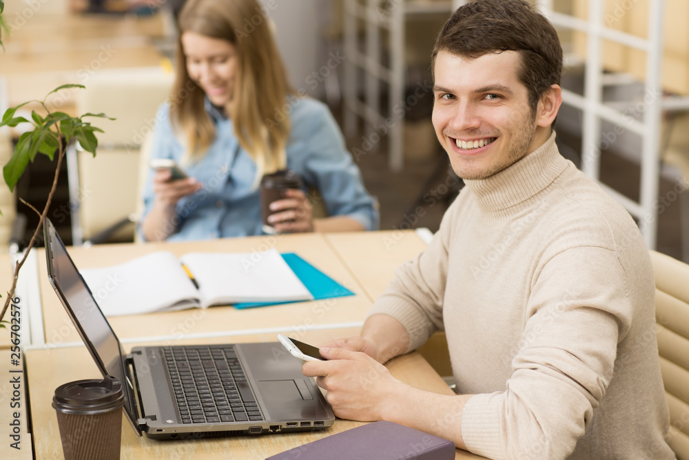 Handsome young cheerful man smiling to the camera using his smart phone while working on the laptop at the office copyspace casual creative designer businesspeople startup technology mobility carrier Handsome young cheerful man smiling to the camera using his smart phone while working on the laptop at the office copyspace casual creative designer businesspeople startup technology mobility carrier