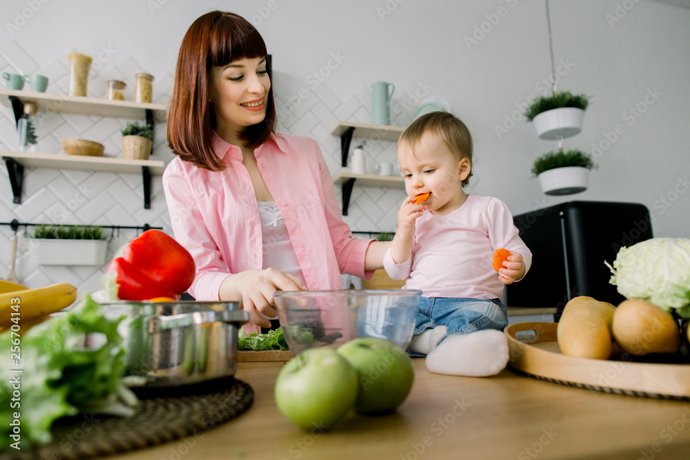 Fototapeta premium Close-up portrait of smiling young mother preparing fresh salad in glass bowl while standing at the table in modern kitchen. Little baby girl eating the carrot. Healthy food concept