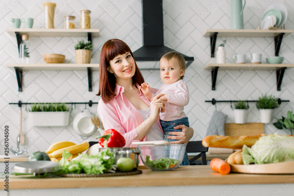 Funny picture of cute little girl with her mom at the table in the kitchen and looking at camera. Young beautiful woman with daughter cooking fresh salad at home. Funny picture of cute little girl with her mom at the table in the kitchen and looking at camera. Young beautiful woman with daughter cooking fresh salad at home.
