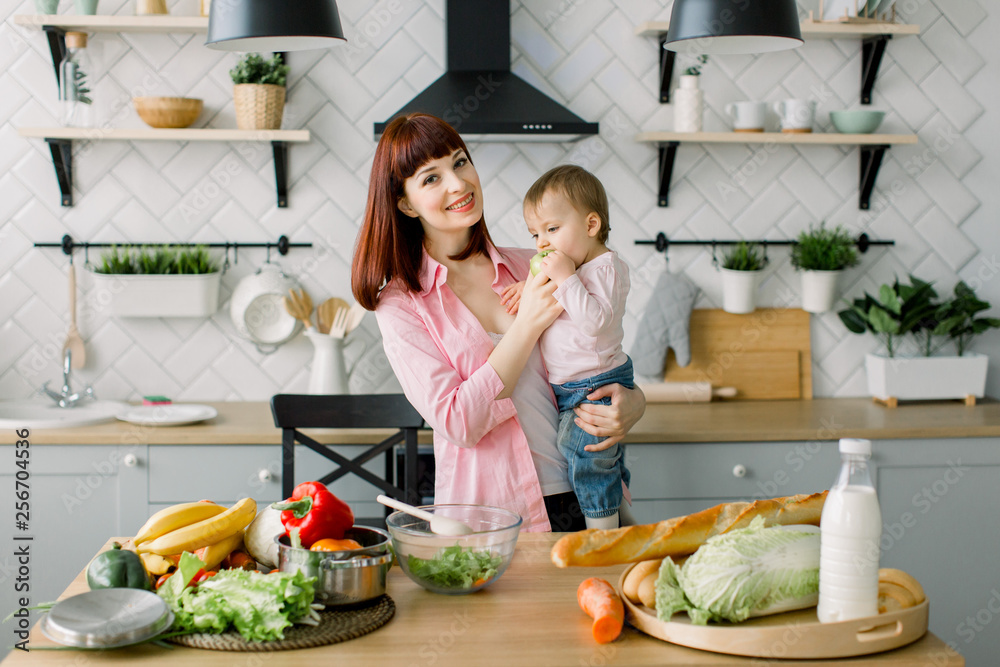 happy smiling mother holding little baby girl eating fresh green apple, standing at the table at home kitchen. Motherhood, family, food, healthy eating, cooking and people concept happy smiling mother holding little baby girl eating fresh green apple, standing at the table at home kitchen. Motherhood, family, food, healthy eating, cooking and people concept